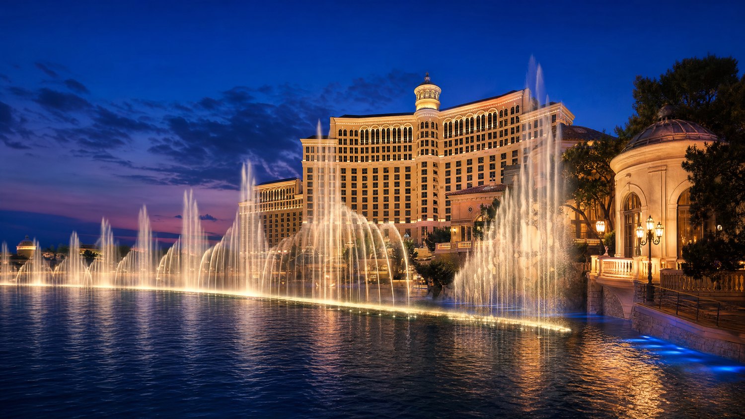 Evening fountain arcs in front of a grand lakeside resort facade.